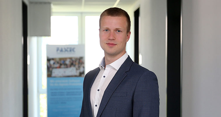 man smiling, standing in a hallway, wearing a suit and white shirt.